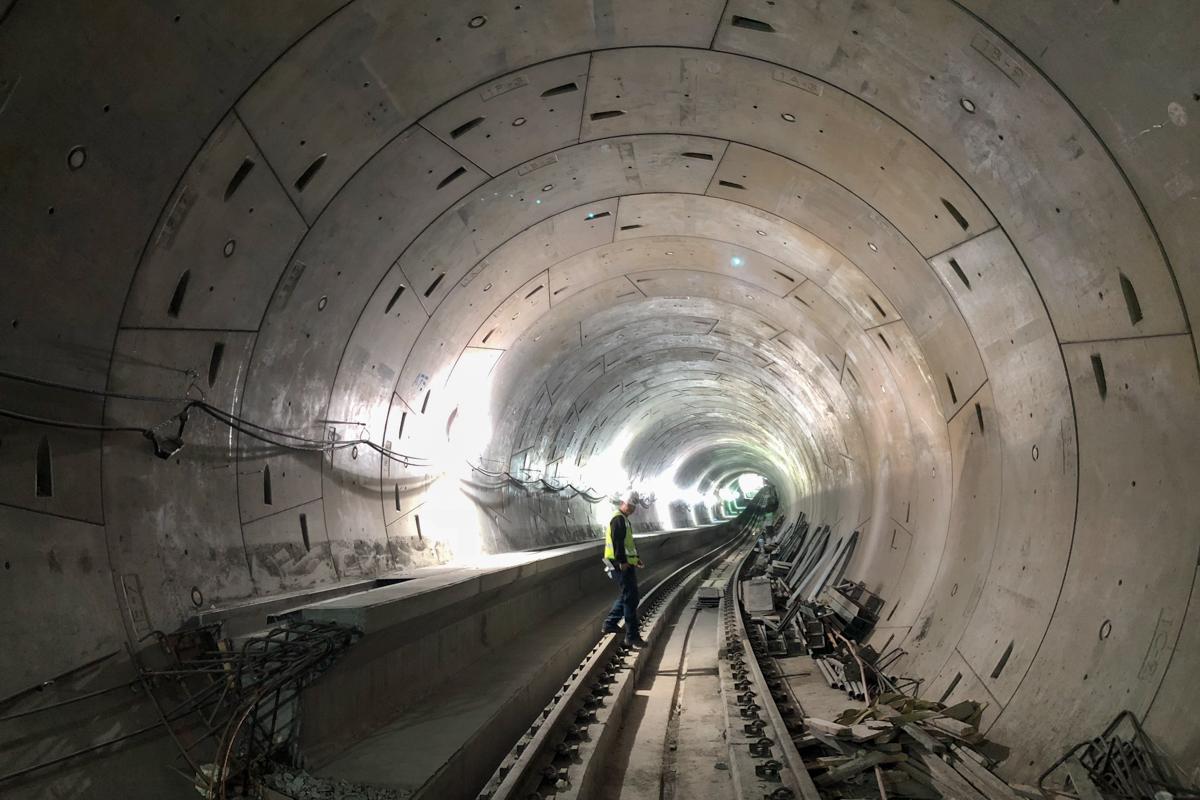Worker in yellow safety vest stands on railroad tracks inside a concrete subway tunnel under construction.