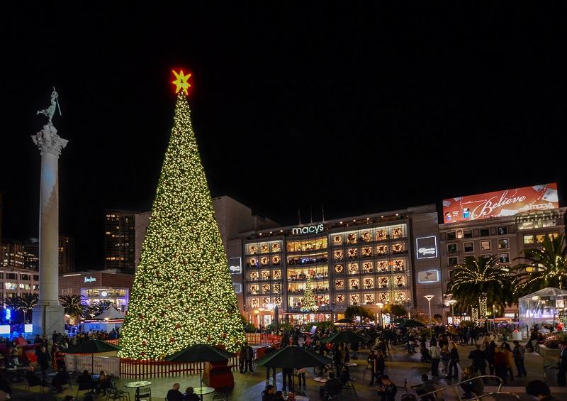 Illuminated Christmas tree with gold lights and star on top at Union Square, surrounded by holiday crowds and decorated storefronts.