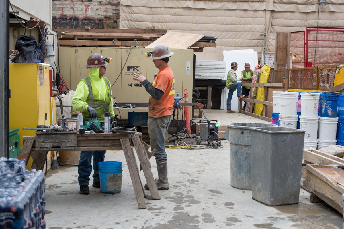 Workers in safety gear operate equipment and machinery in an underground construction work area with concrete structures and storage containers.