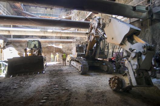 Excavator with grinding wheel and bulldozer inside underground construction headhouse beneath concrete structure.