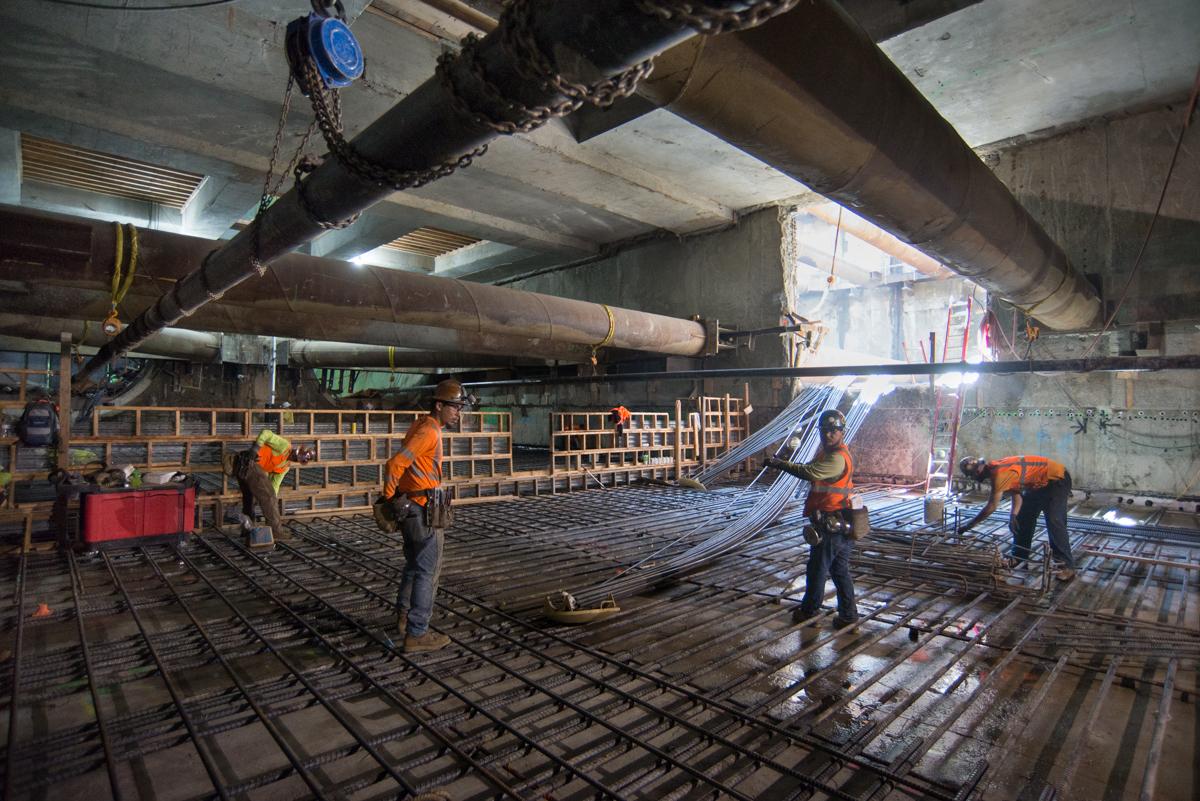 Construction workers in orange vests install reinforcement steel on station platform level beneath concrete beams and overhead bracing.