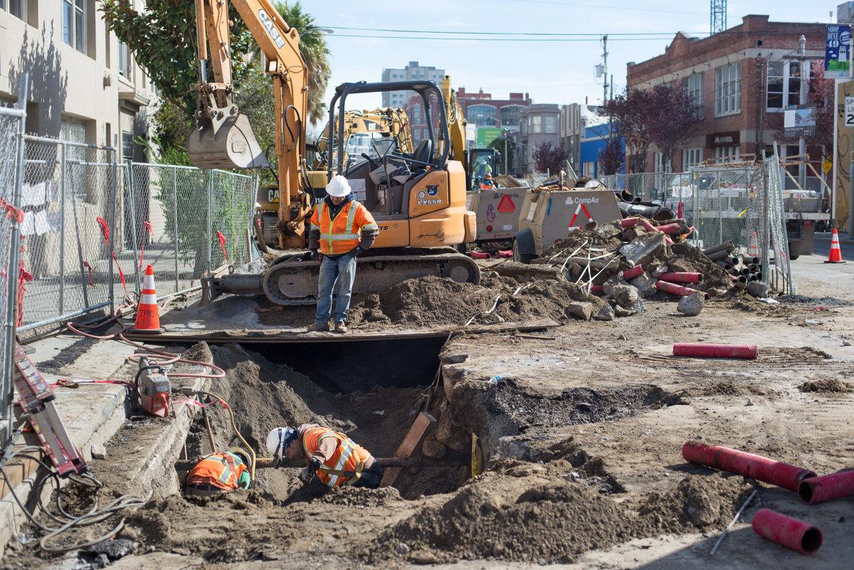 Construction crew operates excavator to dig trench for Central Subway utility installation on city street.