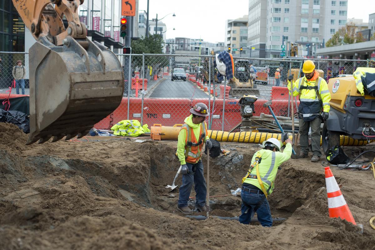 Construction workers in safety vests install utilities in an excavated pit on 4th Street, with heavy equipment and orange barriers visible.