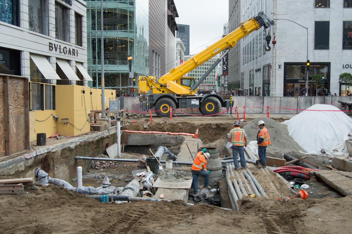Construction site with yellow crane, workers in safety vests, excavated utilities, and temporary barriers on urban street surrounded by buildings.