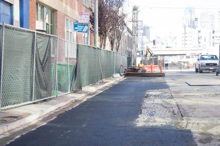Construction site on urban street with green fencing, equipment, and partially restored asphalt near brick building.