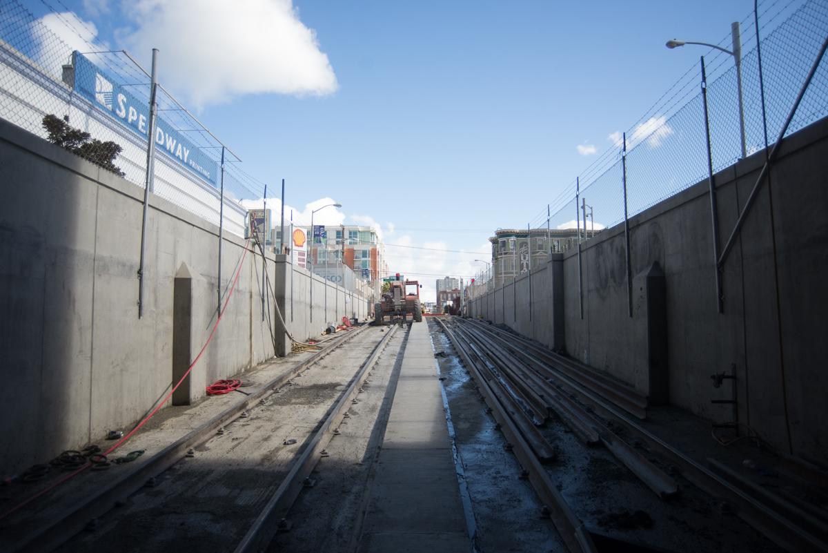 Concrete tunnel walls with steel rails and construction equipment visible along a transit corridor under blue sky.
