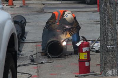 Worker welding large steel pipe section on street with fire extinguisher and traffic cones nearby.
