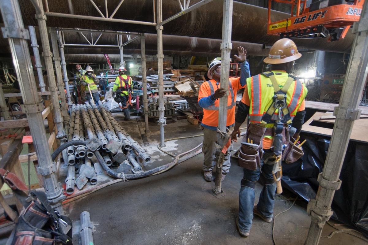 Construction workers in safety gear assemble scaffolding and structural supports inside Chinatown Station.