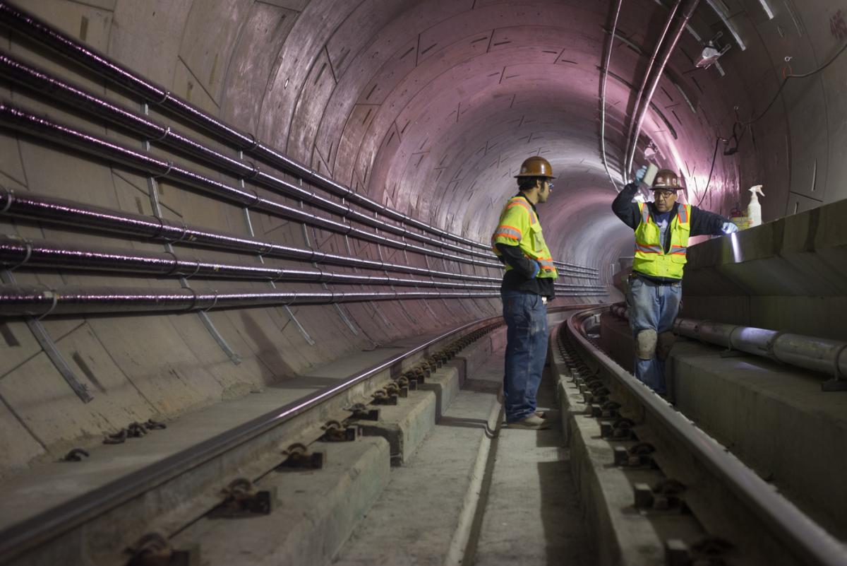 Two workers in hard hats and safety vests inside a tunnel with purple lighting, standing on railroad tracks near concrete walkway.