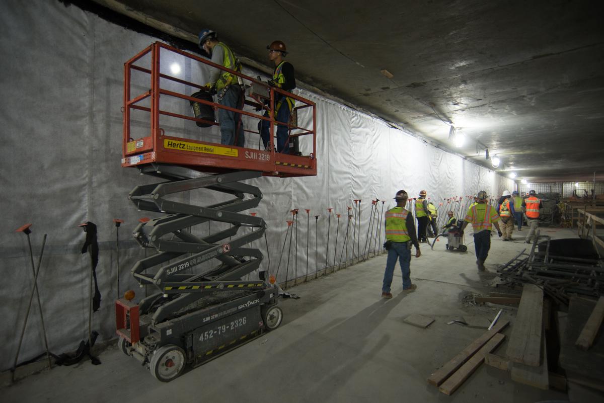 Workers on scissor lift installing waterproofing in underground transit station construction area.