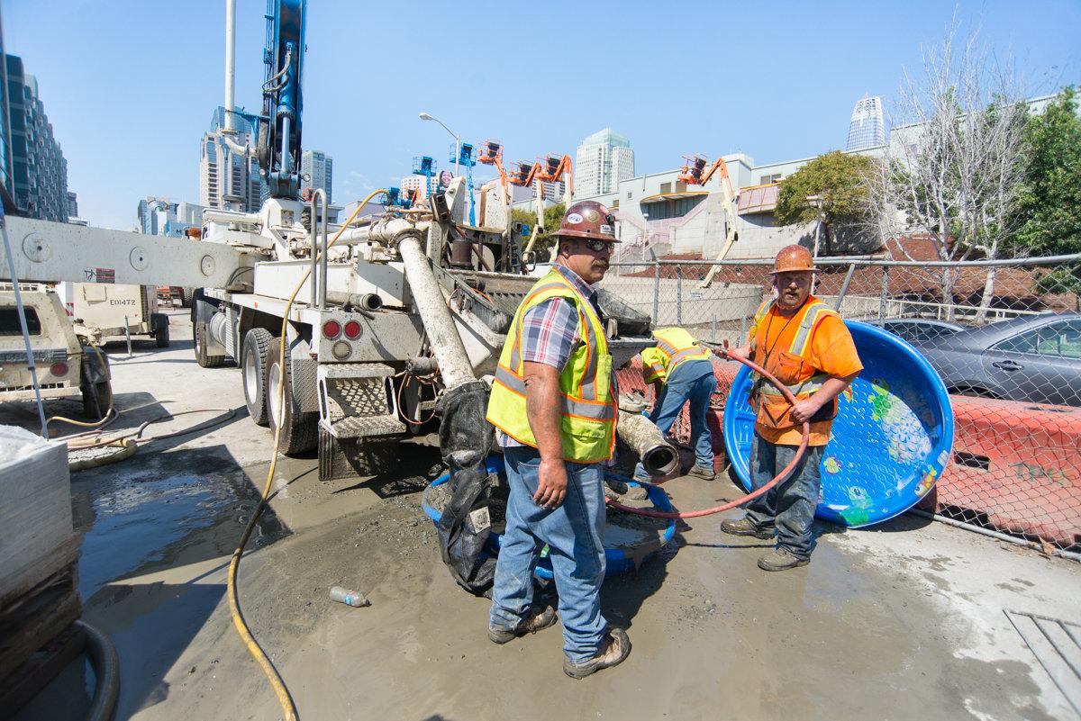Workers operating concrete pumping equipment at a construction site on 4th Street with city skyline in background.