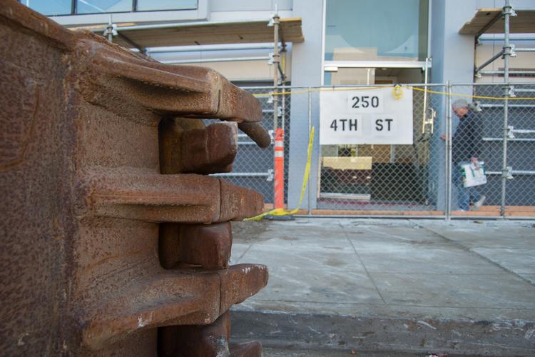 Rusted clamshell bucket teeth at 250 4th Street construction site with chain-link fence gate in background.