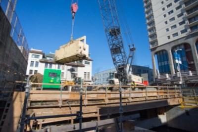 Construction crane lifting yellow steel bucket of excavated material at urban transit site with wooden barriers and multi-story buildings.