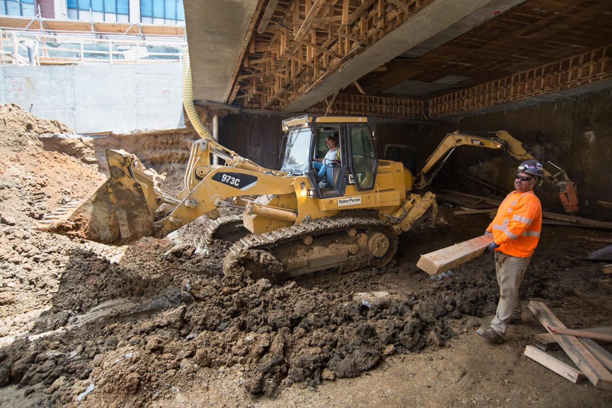 Excavator removing soil under bridge structure at Chinatown Station construction site with worker in orange safety vest