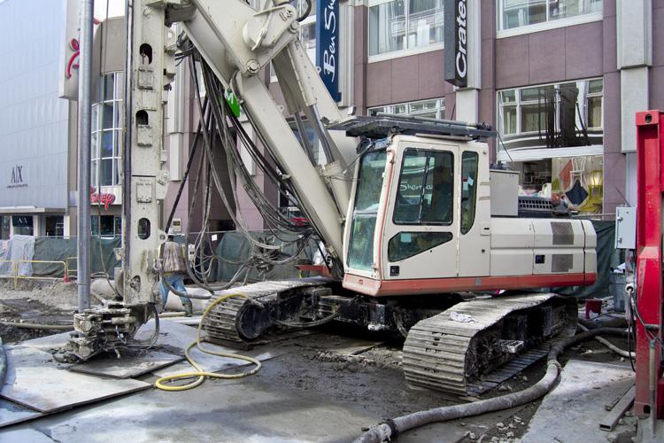 Construction crane with hydraulic arm positioned over excavation site on urban street surrounded by buildings.