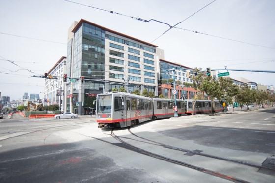 Red and white Muni light rail vehicle on tracks at 4th and King Street with modern buildings in background.