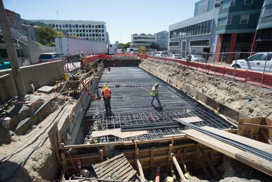 Construction site showing reinforced concrete roof slab being laid with steel rebar, workers in safety gear, and surrounding urban buildings.