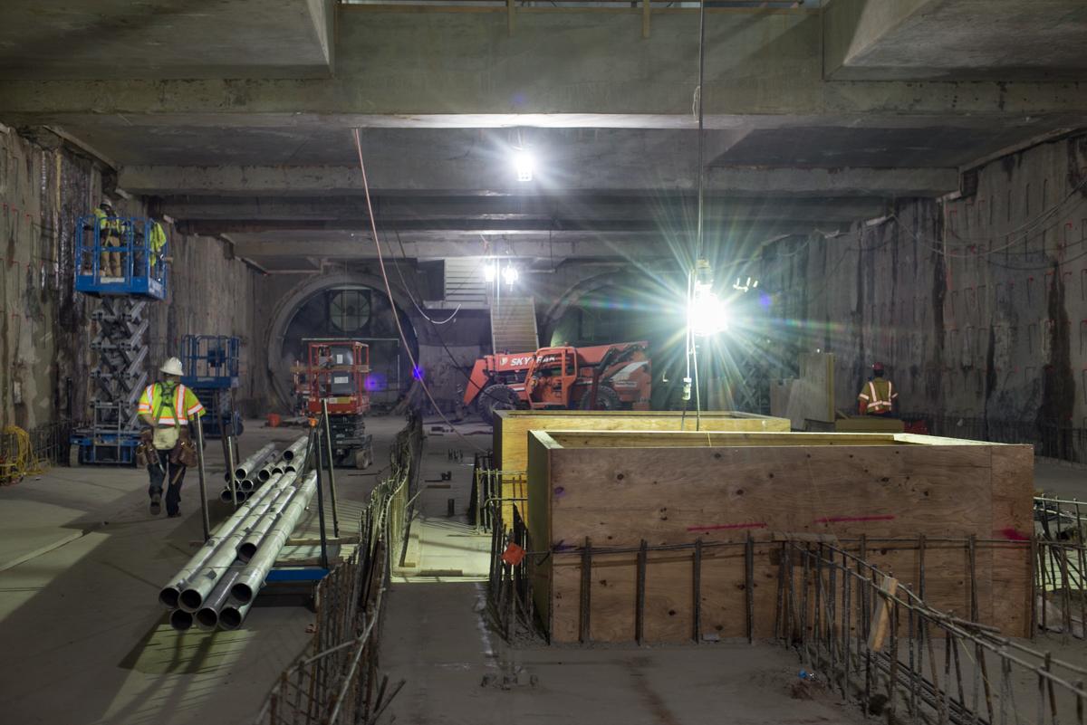 Workers prepare station headhouse interior for rebar installation, with drilling equipment and metal pipes visible under bright work lights.