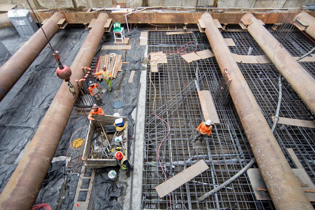 Workers in hard hats install reinforcement rebar on concrete foundation slab during underground transit construction.