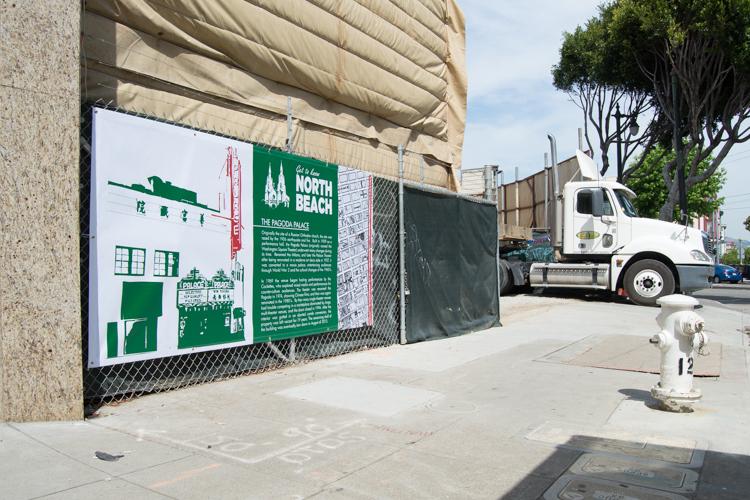 Construction fence with North Beach banner and informational signage along street with white truck parked nearby.
