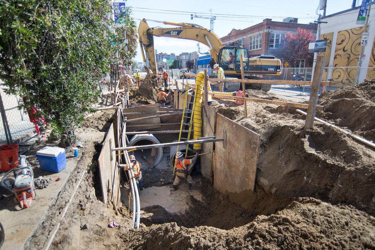 Construction crew working in excavated trench with heavy equipment installing underground utilities on city street.