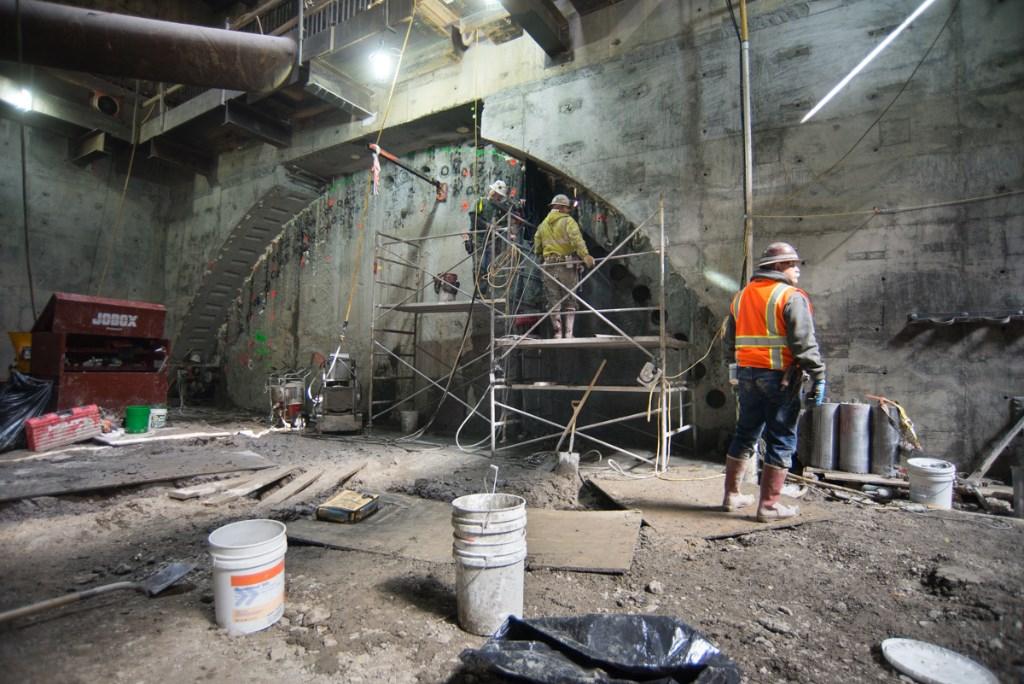 Workers in safety gear perform excavation and concrete work inside an underground transit station during construction.