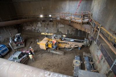 Drill rig operating in underground station excavation with workers and equipment visible in concrete-lined pit.