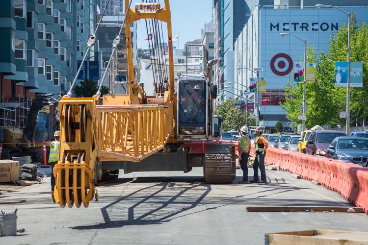 Yellow construction crane with grapple bucket on 4th Street, workers in safety vests nearby, red barriers, urban buildings in background.