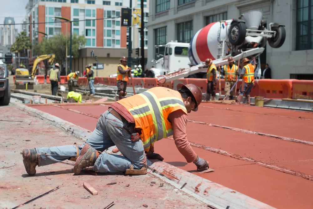Construction worker kneeling on red concrete surface while cement truck pours in background on city street.