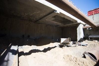 Construction site showing excavated trench with steel support structure and sandy soil under concrete overpass.