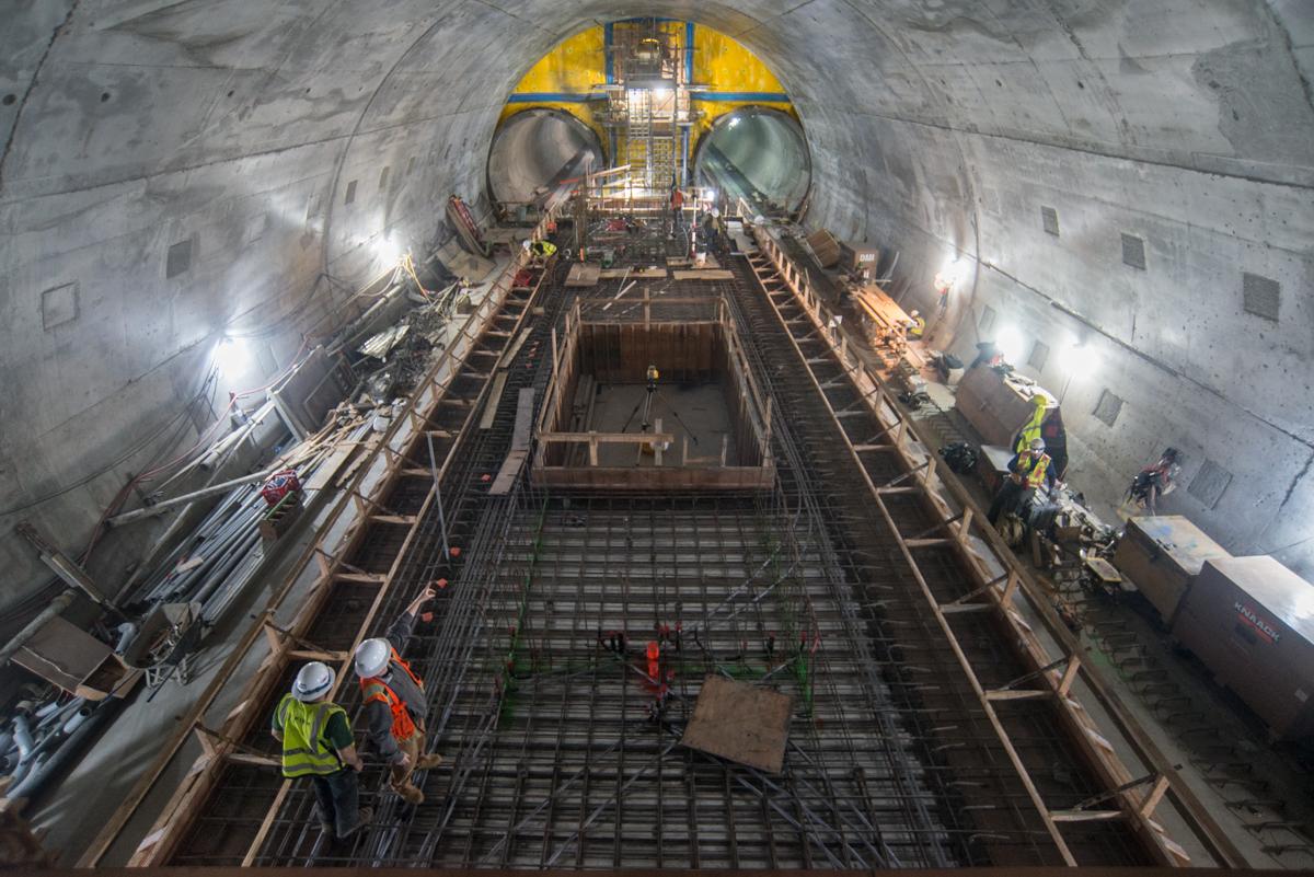 Interior of tunnel under construction with workers, metal rebar framework, and yellow equipment visible overhead.
