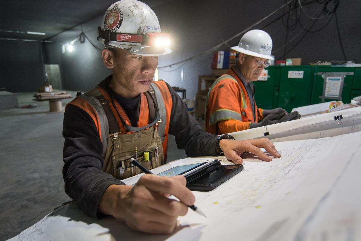 Two construction workers in hard hats and safety vests review blueprints at a table on a mezzanine level.
