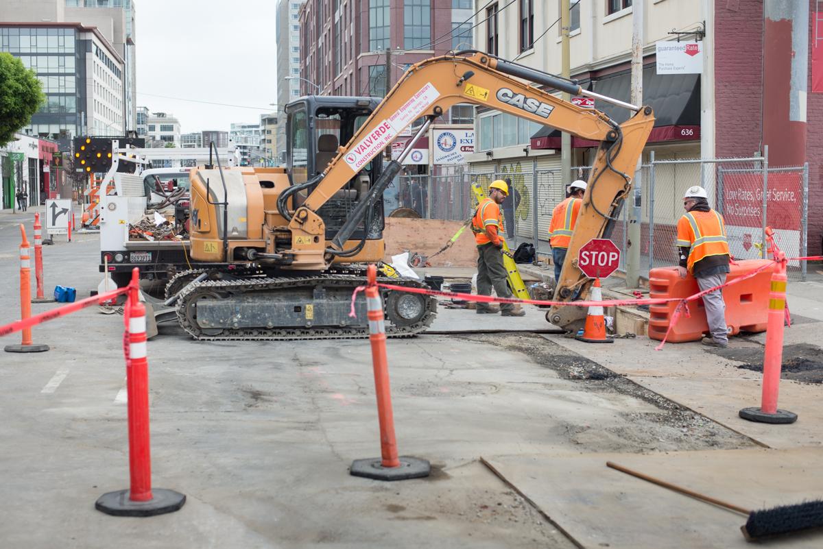 Construction crew operates excavator on 4th Street for utility work, surrounded by safety barriers and traffic cones.
