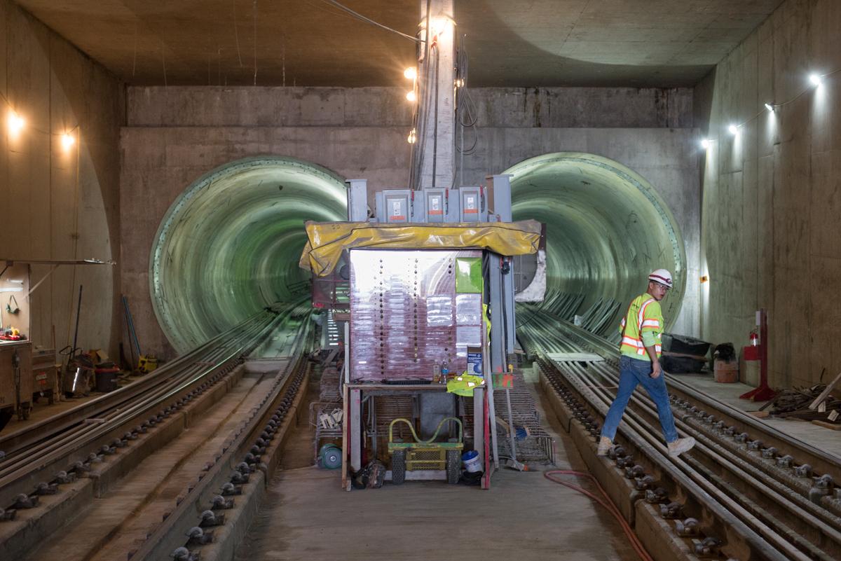 Construction worker in safety gear stands on railroad tracks inside tunnel with two large circular tunnel boring machine openings visible.
