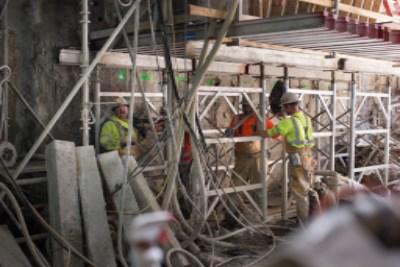 Workers in safety vests on metal scaffolding at underground construction site with excavation equipment visible.