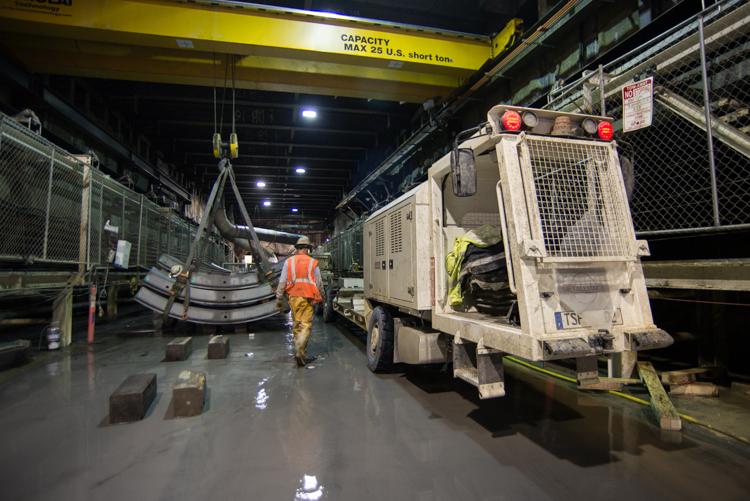 Worker in safety vest guides tunnel segment being lowered by overhead crane into underground launch box facility.