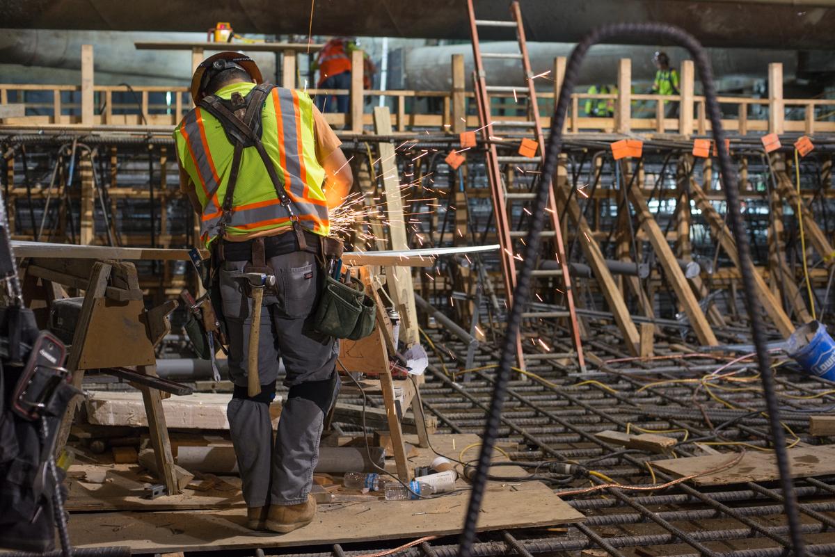 Construction worker in safety gear cutting metal rebar at underground station platform during temporary bracing installation.
