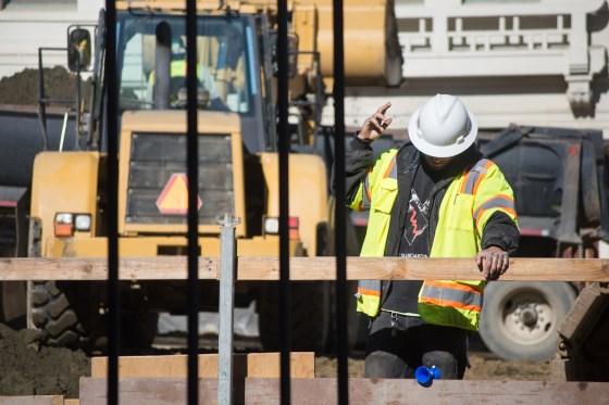 Construction worker in safety vest and hard hat signals near excavation equipment and bucket on Stockton Street.