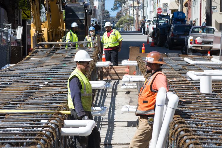 Construction workers in safety vests and hard hats prepare steel reinforcement cages for underground wall construction on a city street.