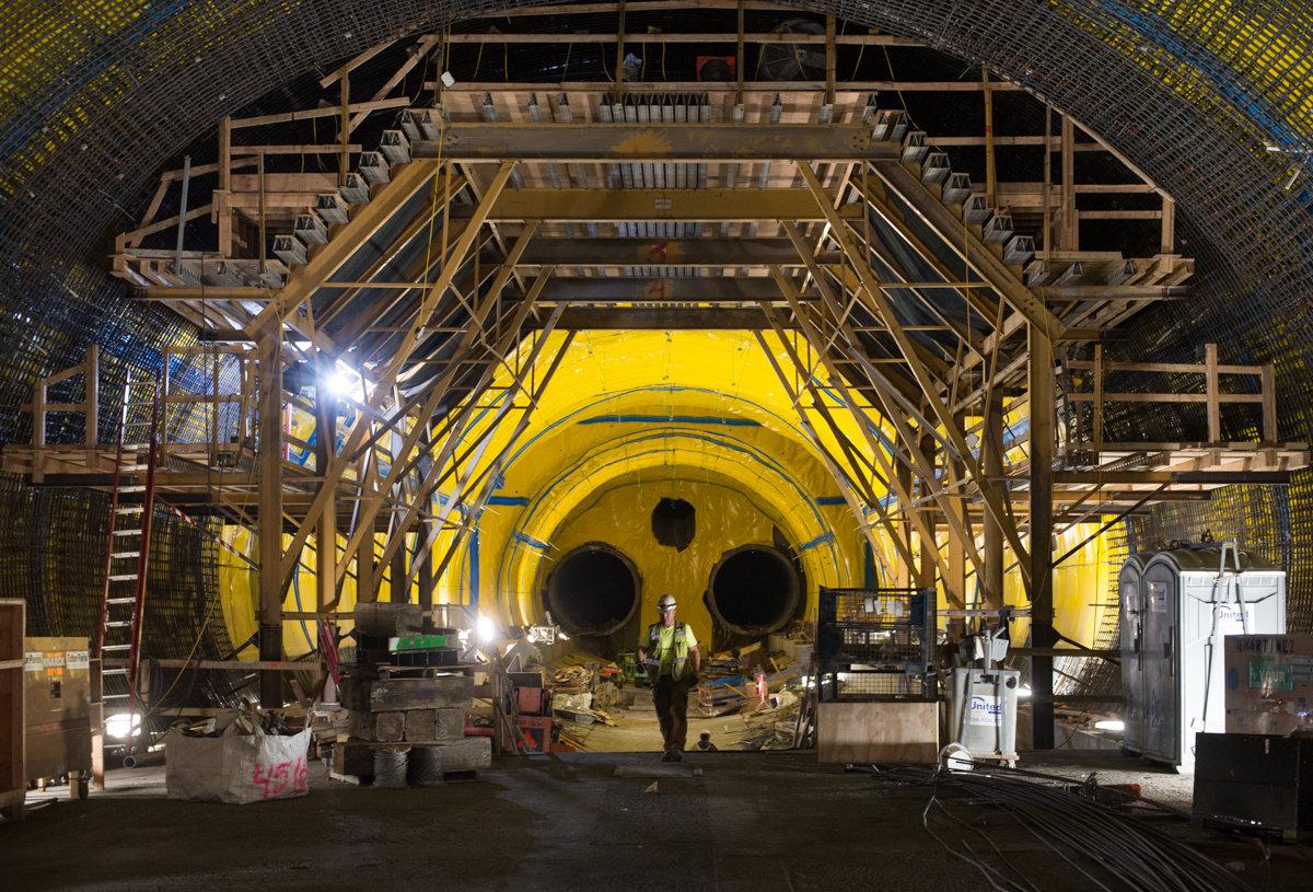 Interior of tunnel under construction with yellow walls, steel reinforcement framework, and two large circular openings visible in distance.