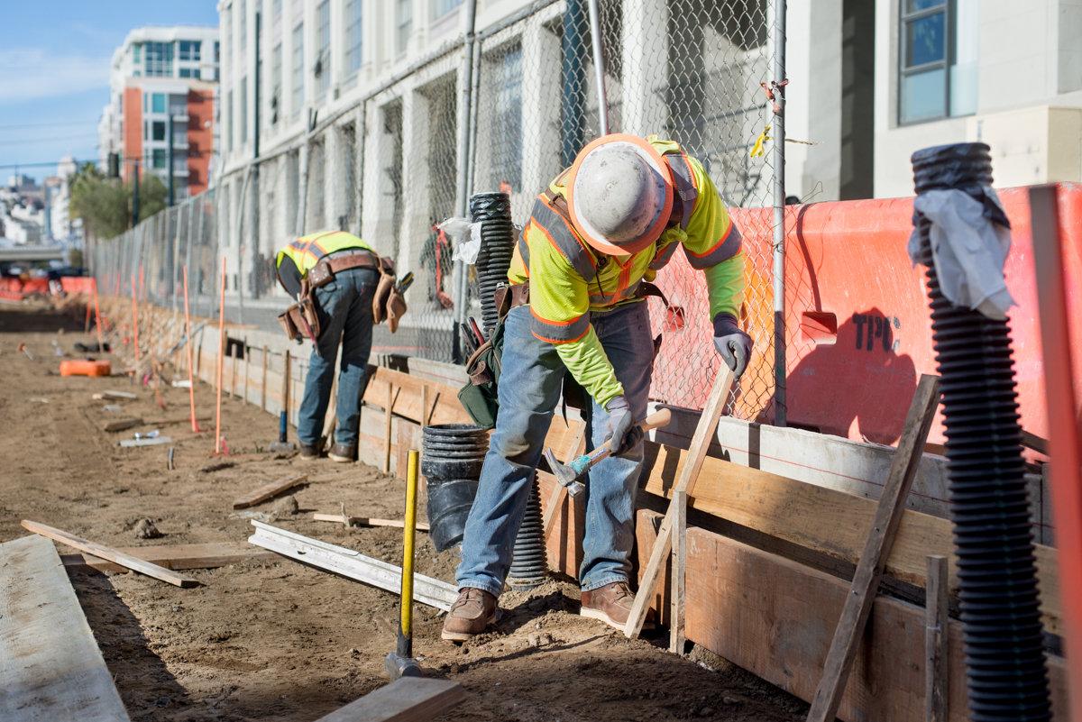 Construction workers in safety vests and hard hats excavating soil at an urban transit project site.