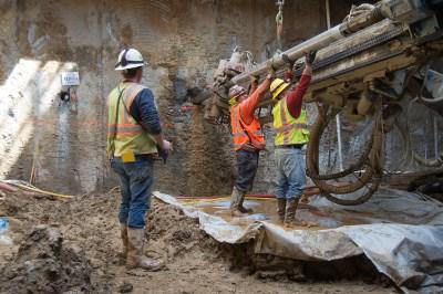 Workers in safety vests operate drilling equipment during dewatering well installation on a city street.