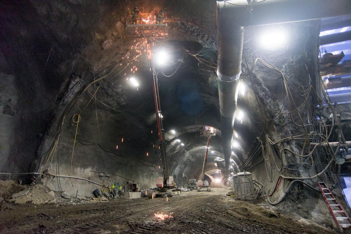 Nighttime tunnel construction with workers in a man basket cutting drill casing segments, sparks flying, heavy equipment and lighting visible.