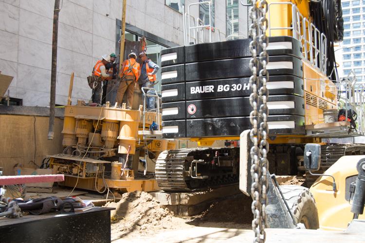 Construction crew operates Bauer BG 30 H drilling rig on urban street with lane closures.