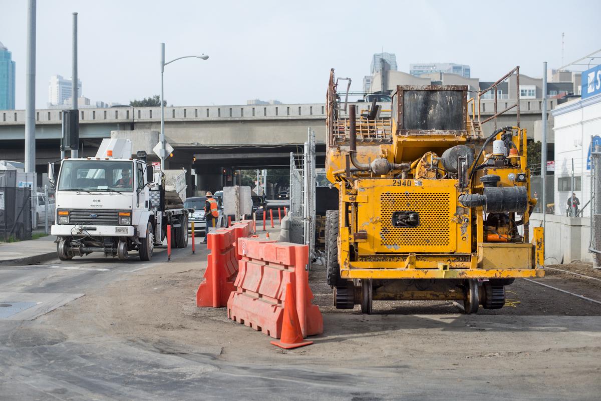 Construction equipment and vehicles staged at transit work site with red safety barriers and elevated highway bridge in background.