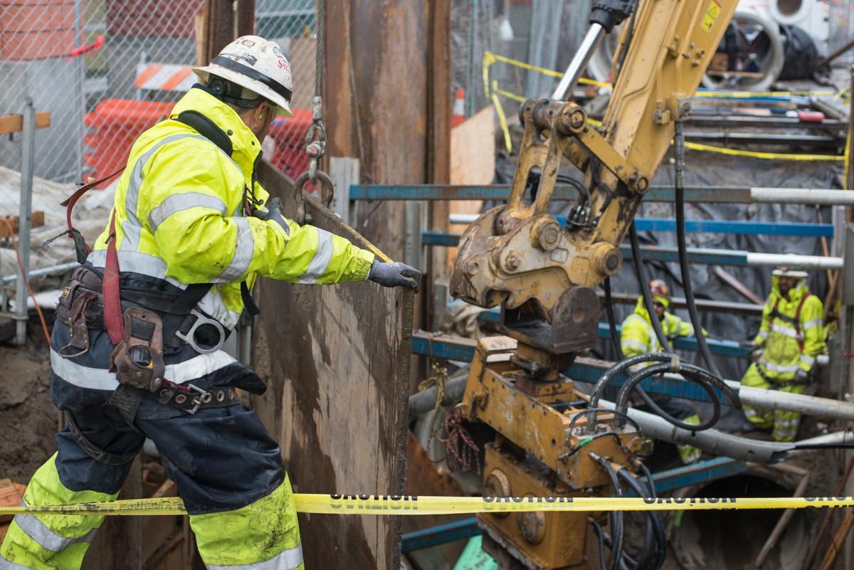 Construction worker guides excavator bucket while installing shoring at 4th and Welsh during Central Subway Project work.