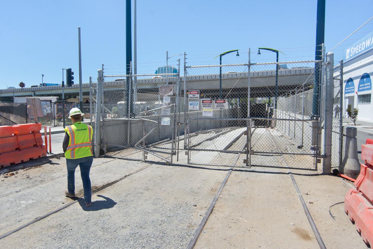 Construction worker in safety vest stands near locked chain-link gate at transit tunnel portal entrance on urban street.