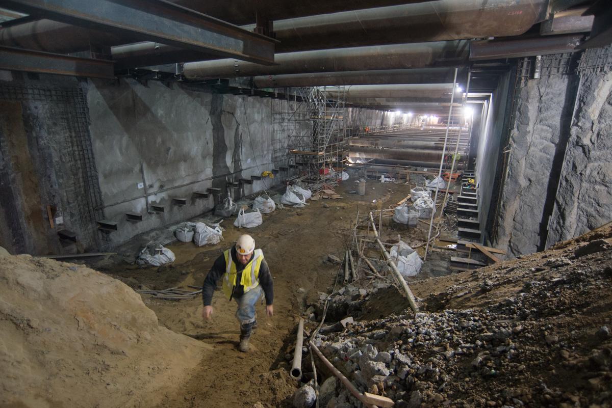 Worker in yellow safety vest walks through underground station construction area with steel beams and temporary bracing.
