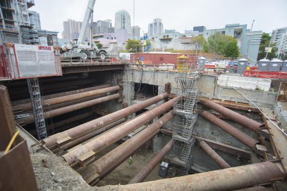Construction pit with large steel pipes and bracing for Yerba Buena/Moscone Station, San Francisco skyline in background.