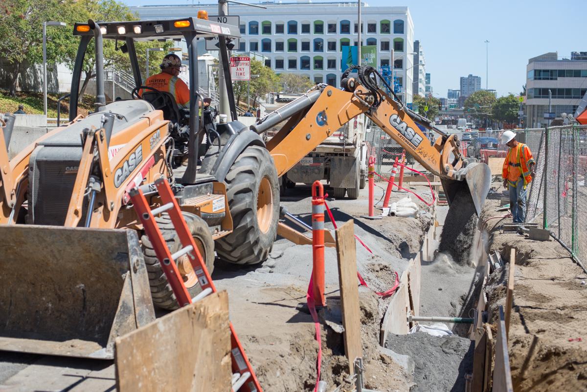 Construction equipment and workers excavating a trench for water line installation at a transit station site.